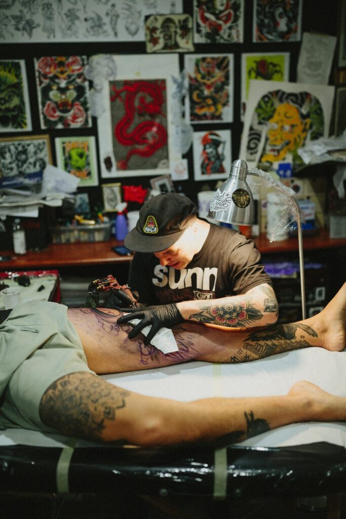 A tattoo artist in a cap tattoos a client's leg