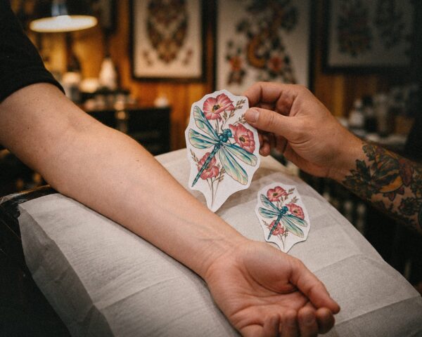 A tattoo artist holds two fine-line flower sketches in different sizes next to a client's forearm